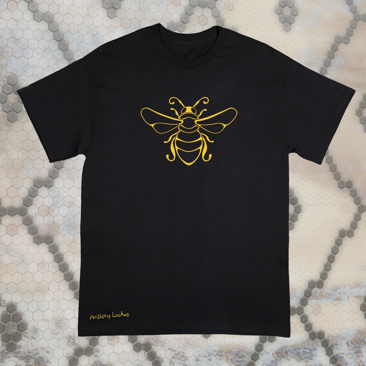 A black short-sleeve Gildan g500 t-shirt with a yellow line art bee graphic on the chest, laid flat on a gray and white hexagonal patterned background. An original hand drawn design by Anthony Lachus with the artists name on the lower left of the shirt. Front view.
