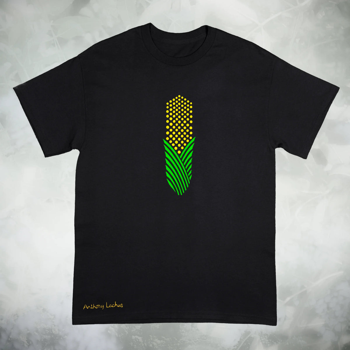 A black short sleeve Gildan g500 crewneck t-shirt featuring a graphic design of a piece of green and yellow corn, laid flat on a textured white background. Original hand drawn artwork by Anthony Lachus. Signature on the lower left of the shirt. Front view.