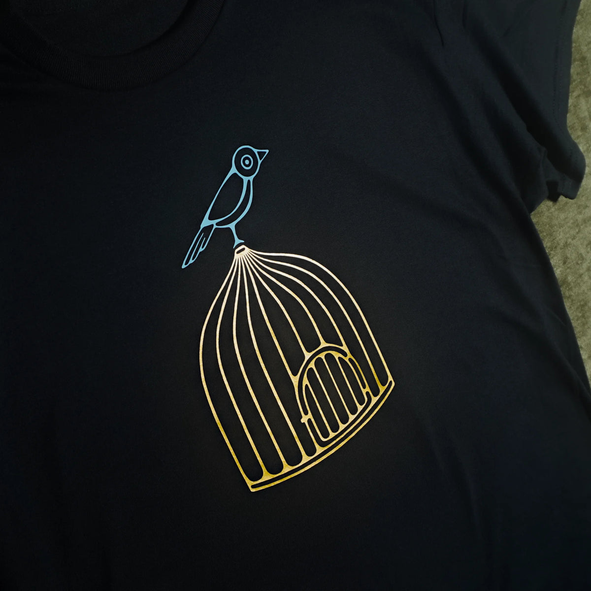 A close-up of a black t-shirt featuring a graphic design of a small bird perched on the corner of a gold-colored birdcage, with the bird outlined in light blue. Alternate view.