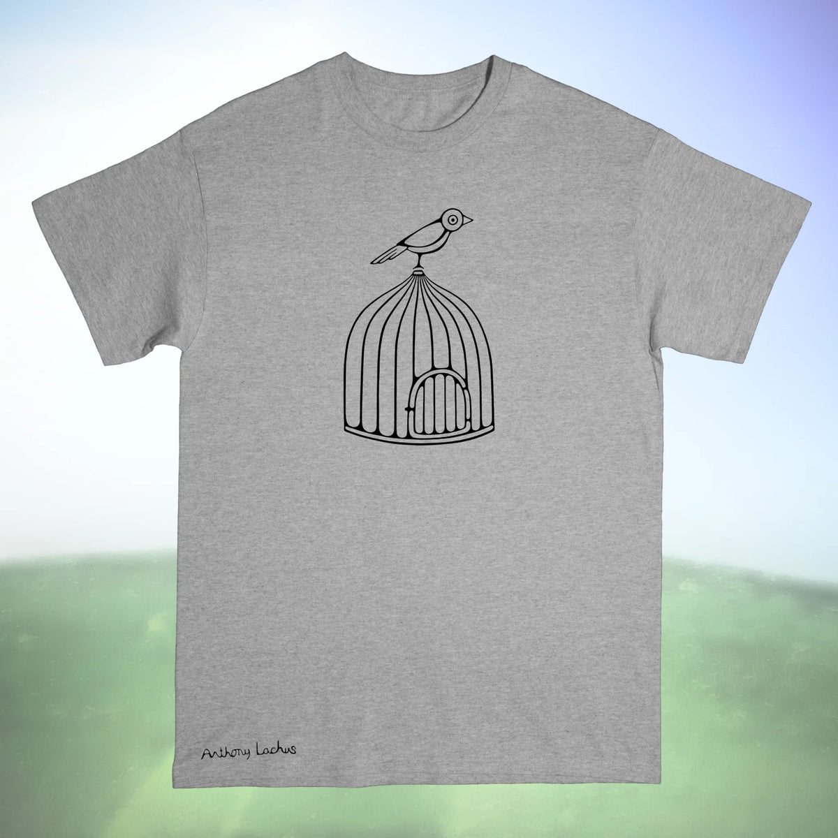 A gray Gildan g500 crewneck t-shirt featuring a graphic design of a black line drawing of a bird perched on a cage, laid flat on a green and white background. An original hand drawn artwork by Anthony Lachus. Front view.