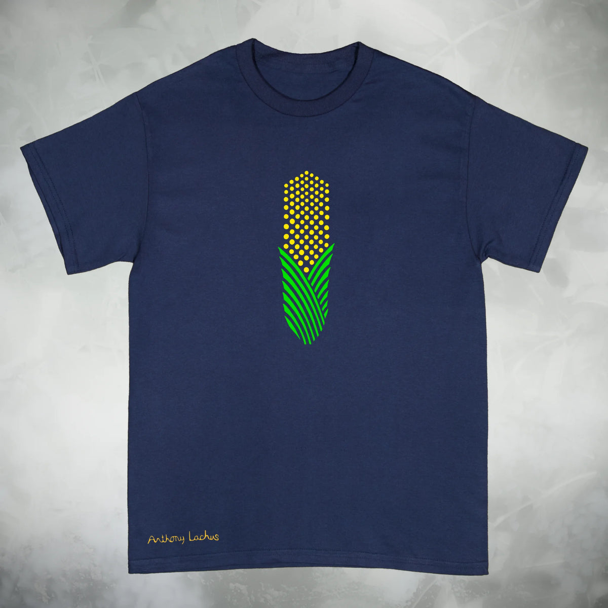A navy blue short sleeve Gildan g500 graphic t-shirt featuring an abstract design of yellow and green corn, laid flat on a textured light gray background. Original hand drawn artwork by Anthony Lachus. Signature on lower left. Front view.
