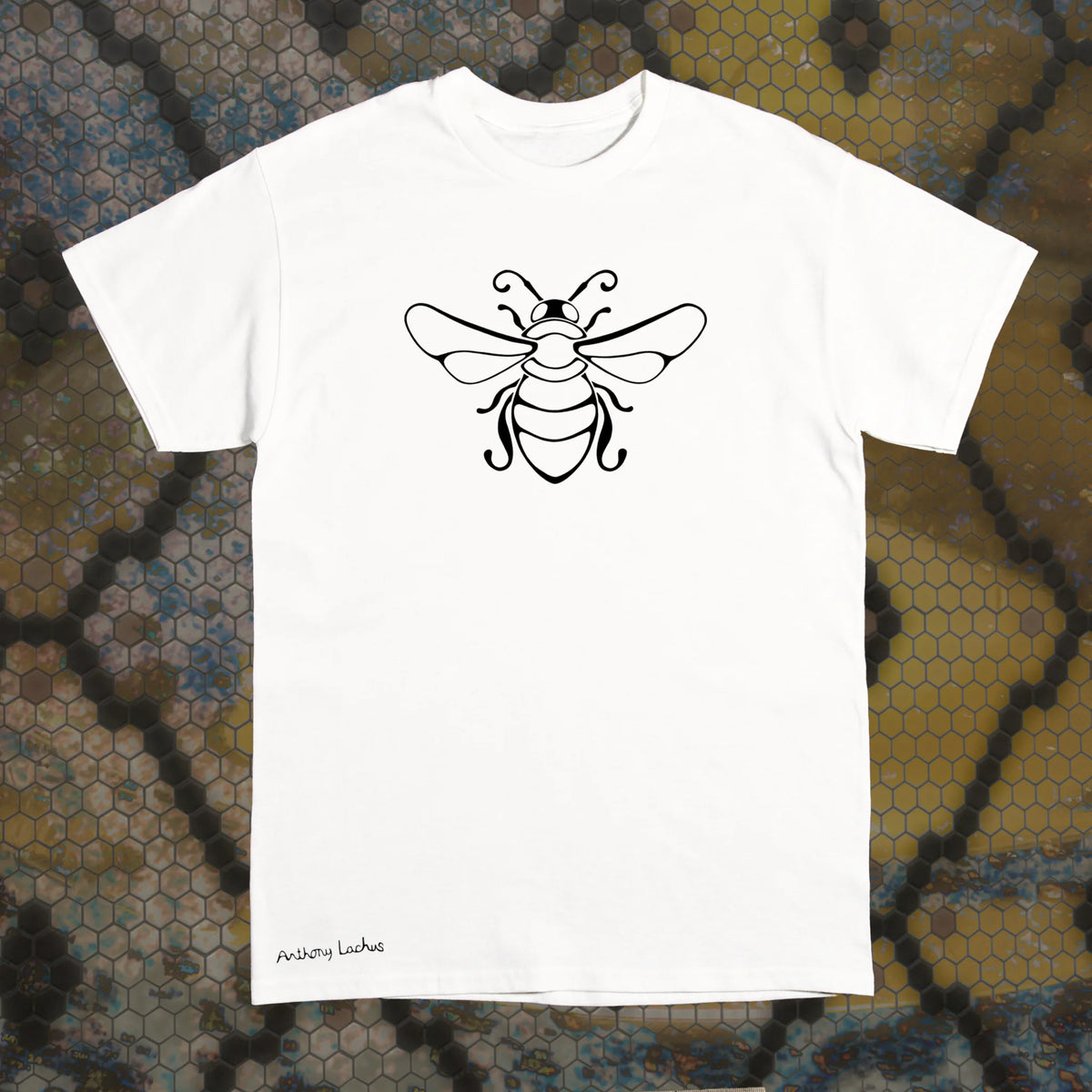 A white short-sleeve Gildan g500 crewneck t-shirt with a black line art bee graphic on the chest, laid flat on a black and yellow hexagonal patterned background. An original hand drawn design by Anthony Lachus with the artists name on the lower left of the shirt. Front view.